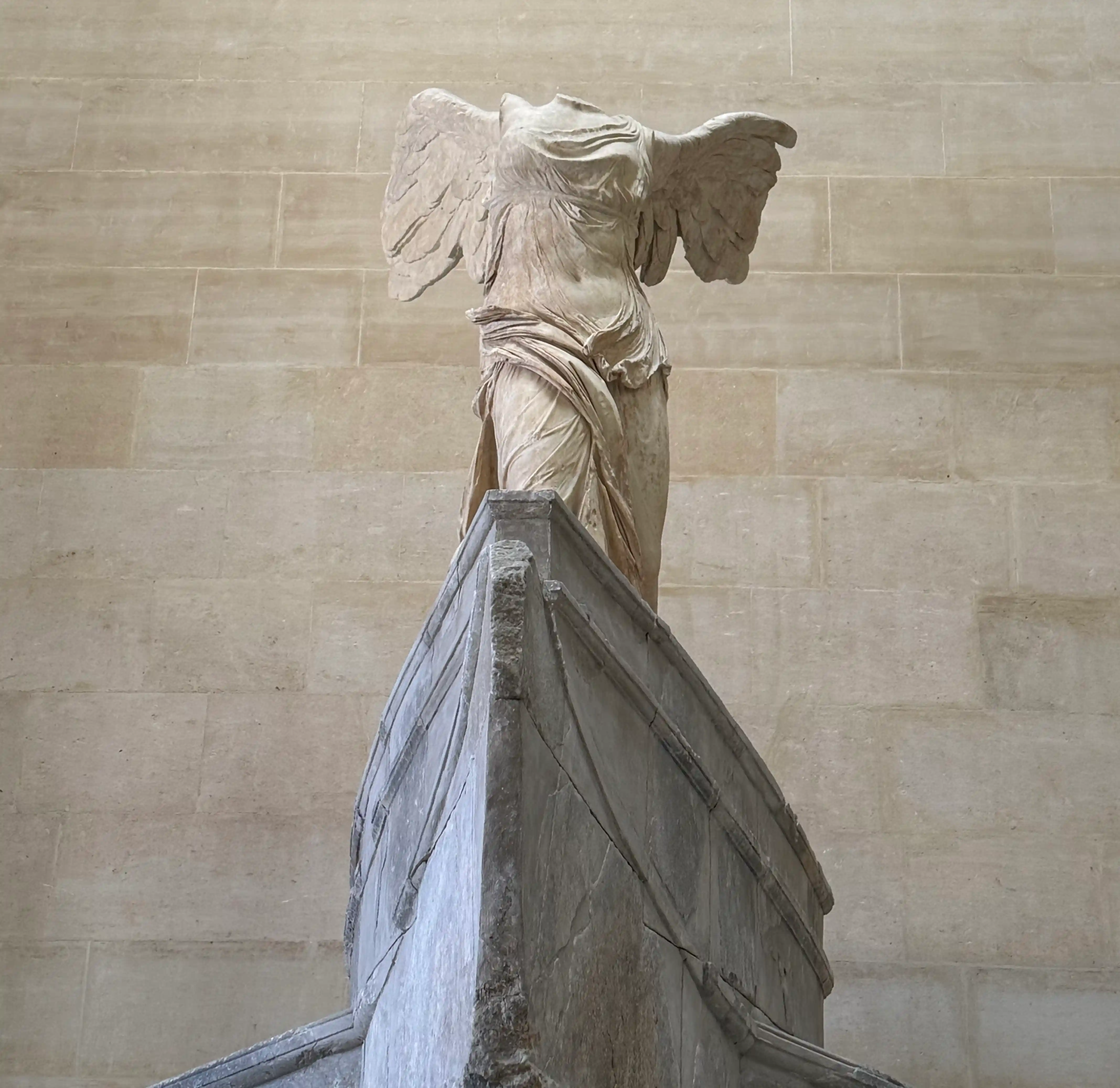 A winged marble statue in front of a tan colored wall.